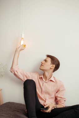 Striptiz tişörtlü ve siyah kotlu genç çekici bir adam içeride oturuyor ve ampulle oynuyor. Düşünceleri, fikirleri, analizleri, yansımaları kavra.