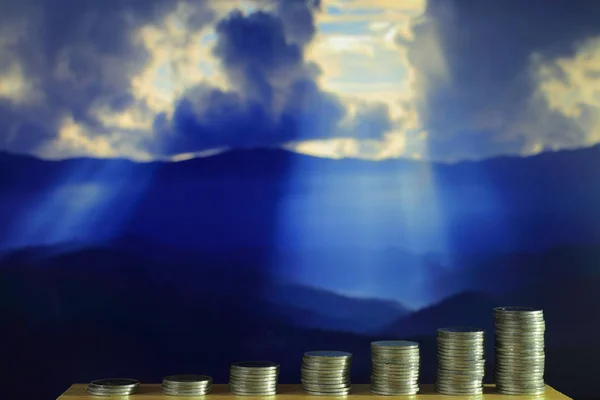 funding for environmental conservation.  pile of coins in increasing chart shape in front of dramatic god light beam from bursting cloud over mountain in the background.