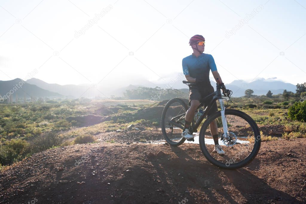 backlit image of a mountain biker and his bike on top of a mound with a mountain range in the distance