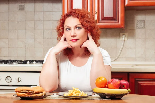 Big woman eat fast food. Red hair fat girl with burger, potato and fruit. Unhealthy food concept with plus size female on kitchen