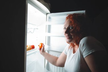 Büyük kadın meyve ye. Kızıl saçlı şişman kız buzdolabı arıyorum. Sağlıksız ve sağlıklı gıda kavramı ile artı boyutu kadın mutfak