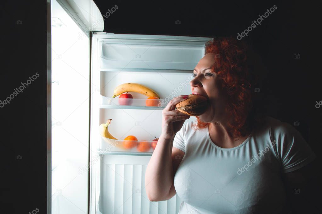Big woman eat fast food. Red hair fat girl looking inside refrigerator with burger. Unhealthy and healthy food concept with plus size female on kitchen