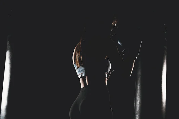 Woman with black boxing wraps and boxing gloves on hands boxing in dark with punching bag. Boxer girl silhouette