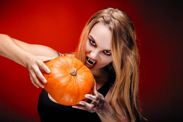 Halloween girl vampire with red eyes red lips and dark red background. Scary woman trick or treat time, halloween concept. Female makeup for holiday with pumpkin in hands
