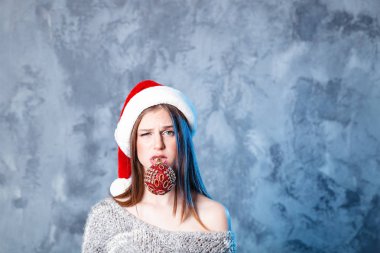 Mutlu Noeller ve mutlu yeni yıl! Sevimli kız ile top oyuncak ağız içinde çok şaşkın görünüyor. Gri arka plan üzerinde yakın portre. Kız santa şapka ve kazak