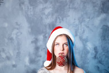 Mutlu Noeller ve mutlu yeni yıl! Sevimli kız ile top oyuncak ağız içinde çok şaşkın görünüyor. Gri arka plan üzerinde yakın portre. Kız santa şapka ve kazak
