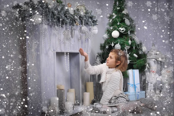 a girl in bright clothes stands near a gray fireplace and a green Christmas tree