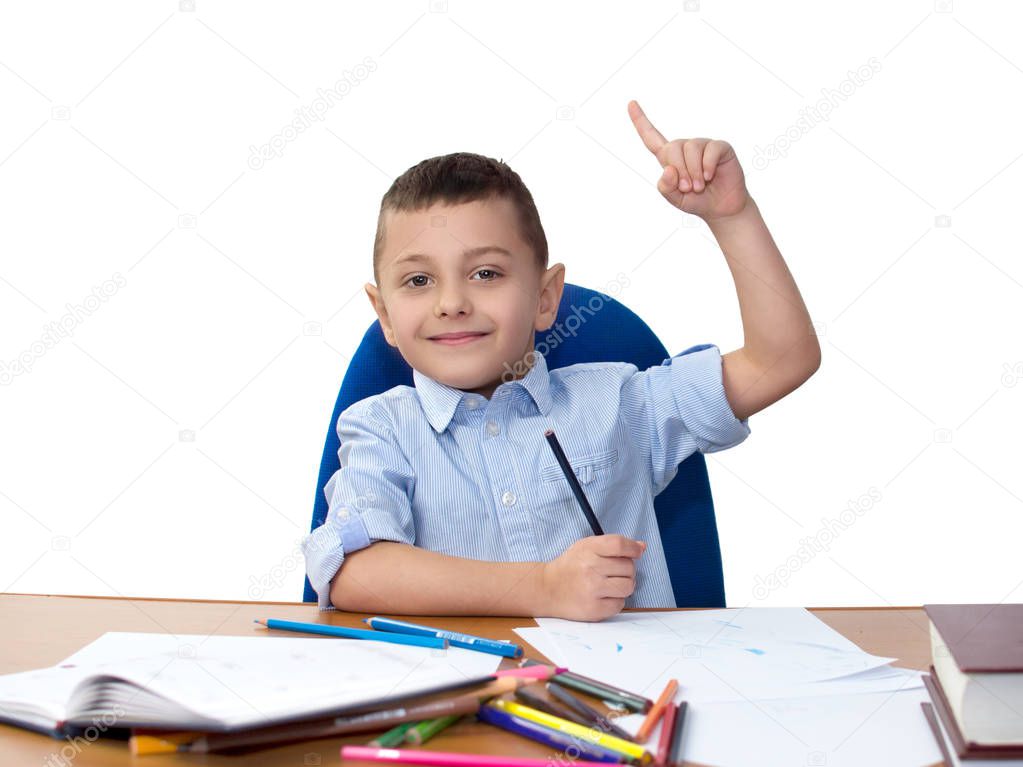 boy student sits on a chair behind a desk, draws with colored pencils and shows his index finger upwards, isolated on white background