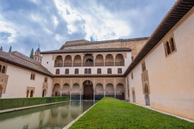 La Alhambra, İspanya - Patio de los Arrayanes 'in uzun yansıtan havuzu, çalıları ve parçalı bulutlu gökyüzünün altındaki Fas kemerleriyle sakin bir avlu manzarası.