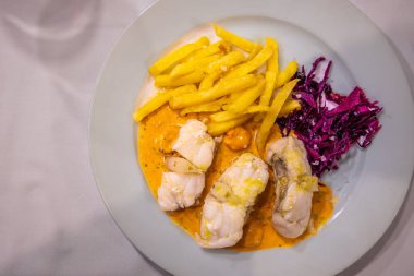 Antequera, İspanya - 12 Mart 2025: Portakal soslu, patates kızartmalı ve lahanalı bir tabak beyaz balık filetosu. Zeytin bahçesindeki bir kır lokantasında hizmet vermiş..