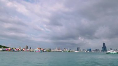 Ferry crosses Kaohsiung Harbor in Taiwan with a backdrop of modern city skyline. Calm water and cloudy sky frame this busy coastal transit scene.
