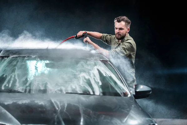 A man with a beard or car washer washes a gray car with a high-pressure apparatus at night in a car wash