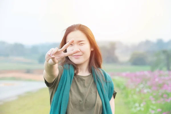 Porträtt av ung asiatisk kvinna tittar på kamera med leende och visar fred tecken med fingrar med Cosmos-fältet i bakgrunden. — Stockfoto