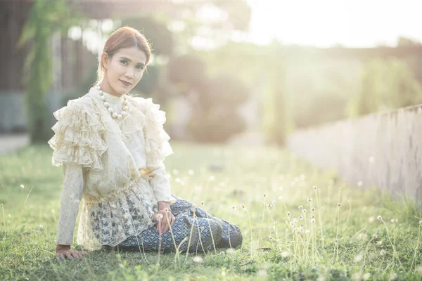 Mujer asiática novia en traje tradicional tailandés sentarse en la hierba al aire libre en una mañana que rodea por la luz del sol de oro y flor de hierba . — Foto de Stock