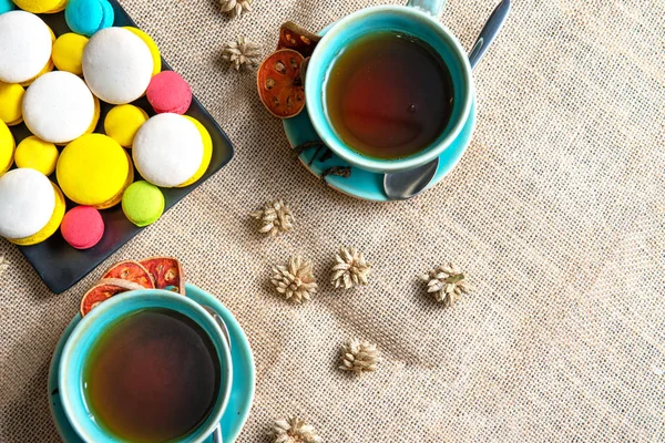 Cup of hot herb black tea on sag background with some fresh colorful macaroon on black plate,tea time concept. — Stock Photo, Image