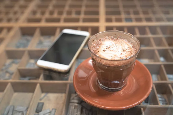 Glass of hot mocha coffee on glass floor with smart mobile phone on table in background. — Stock Photo, Image