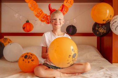 Şeytani boynuzlu sarışın çocuk Cadılar Bayramı yatak odasında bir balon tutuyor balon parlarken boynuzlar parlıyor Cadılar Bayramı yatak odasını şenlikli ve büyüleyici yapıyor. Yüksek kalite fotoğraf