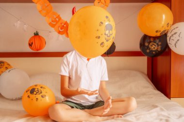 Balonla süslü bir yatak odasında poz veren yakışıklı çocuk şeytan boynuzlarıyla gülümserken balon renk katıyor ve dekore edilmiş yatak odası Cadılar Bayramı fotoğrafları için mükemmel oluyor. Yüksek kalite fotoğraf