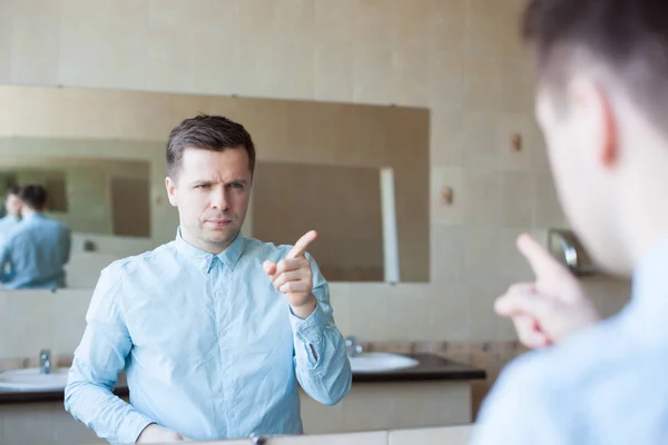 Молодий чоловік у блакитній сорочці налаштовує волосся перед дзеркалом у туалеті. Він турбується і готується до зустрічі — стокове фото