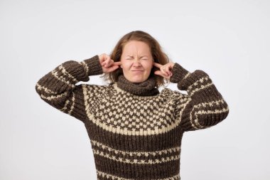 Kulakları tıkalı ve gürültüden rahatsız yüzü çatık sıcak kahverengi kazaklı genç erkek. Yüksek bir ses ve gürültü yorgun sakin rahat yerde kalmak istiyor