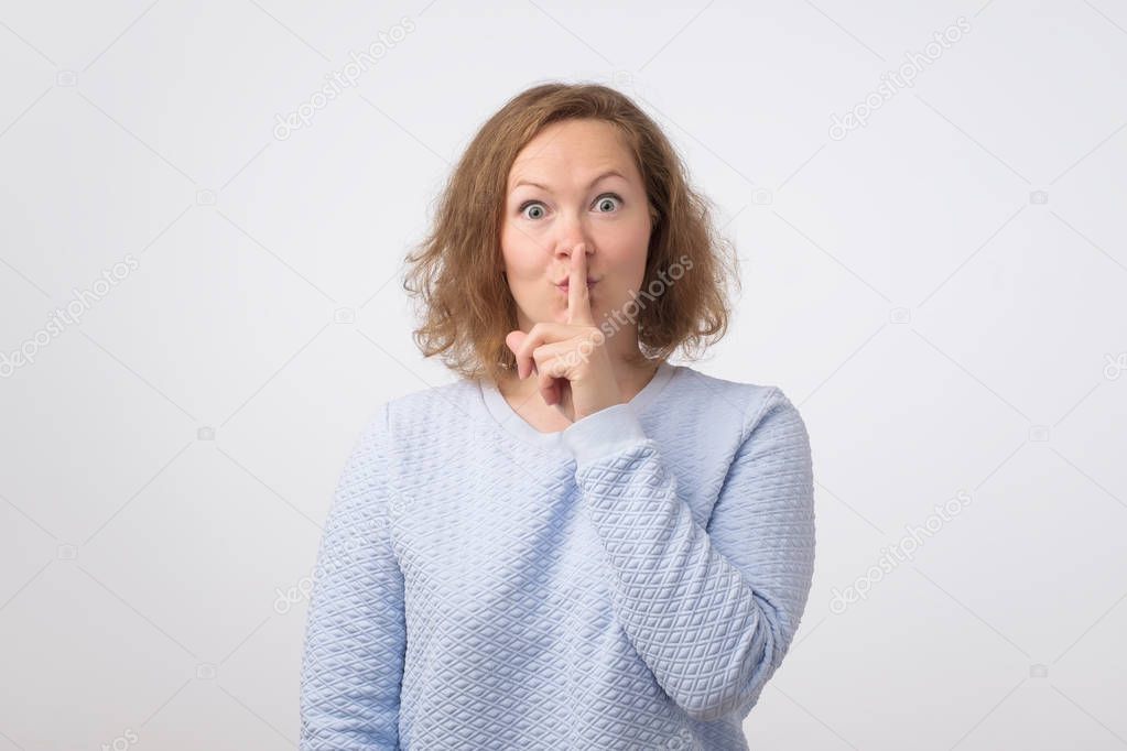 Mature woman in blue sweater making silence gesture with forefinger, isolated on white background