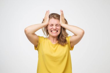 Korkunç başım ağrıyor. Sinir bozucu Avrupa Genç kadın başını elleri ile dokunarak ve gri arka plan karşı dururken acı dolu surat sarı t'shirt içinde