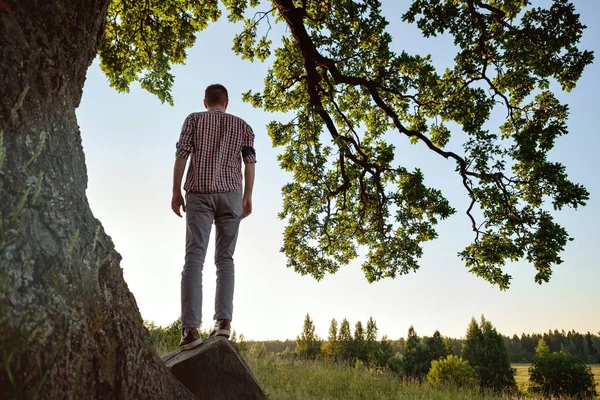 Un hombre con camisa a cuadros de pie cerca del árbol y mirando a un lado en el valle . — Foto de Stock
