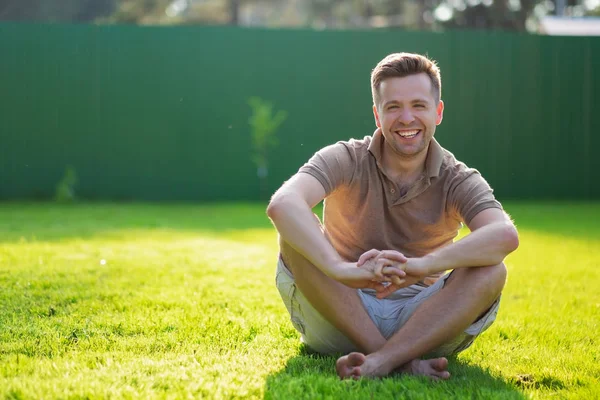 Молодой счастливый человек улыбается в камеру, сидя на зеленой траве — стоковое фото