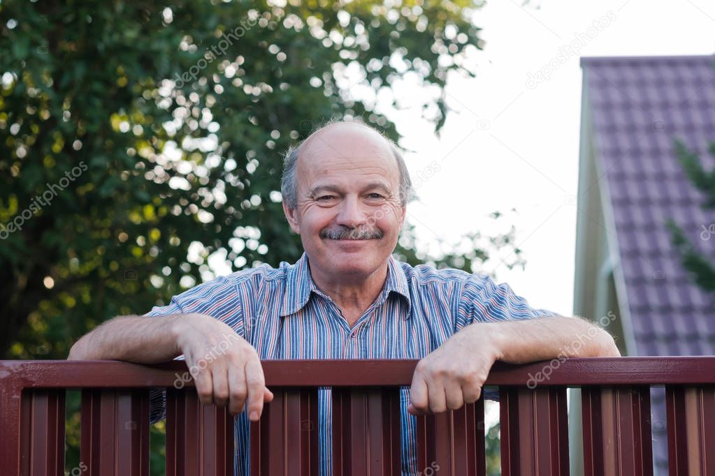 Portrait of a happy handsome mature man standing leaning on the red metal fence. He is waiting for guests to his small country house.