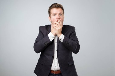 Ağzını el ile gri duvar izole dururken kapsayan takım elbiseli sürpriz genç erkek portresi. Bu haber inanamıyorum..