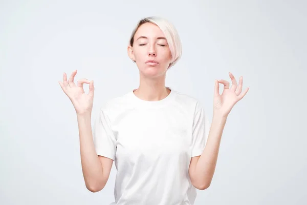 Joven mujer sonriente con peinado corto de color meditando, sosteniendo sus manos en gesto de yoga, sintiendo calma —  Fotos de Stock