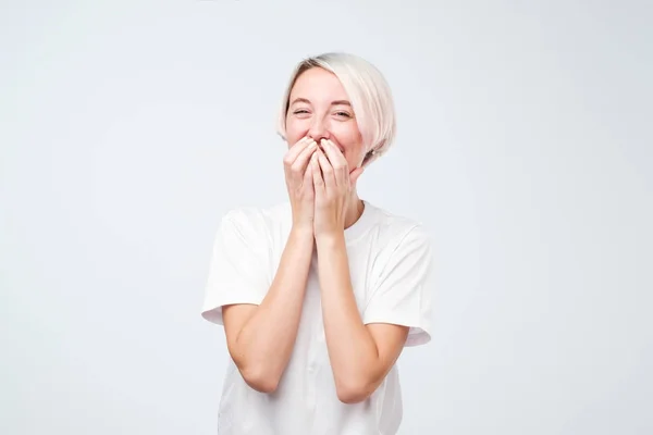 Добре виглядає молода жінка з фарбованим волоссям у білій футболці радісно горить, покриває рот, як намагається зупинити сміх — стокове фото