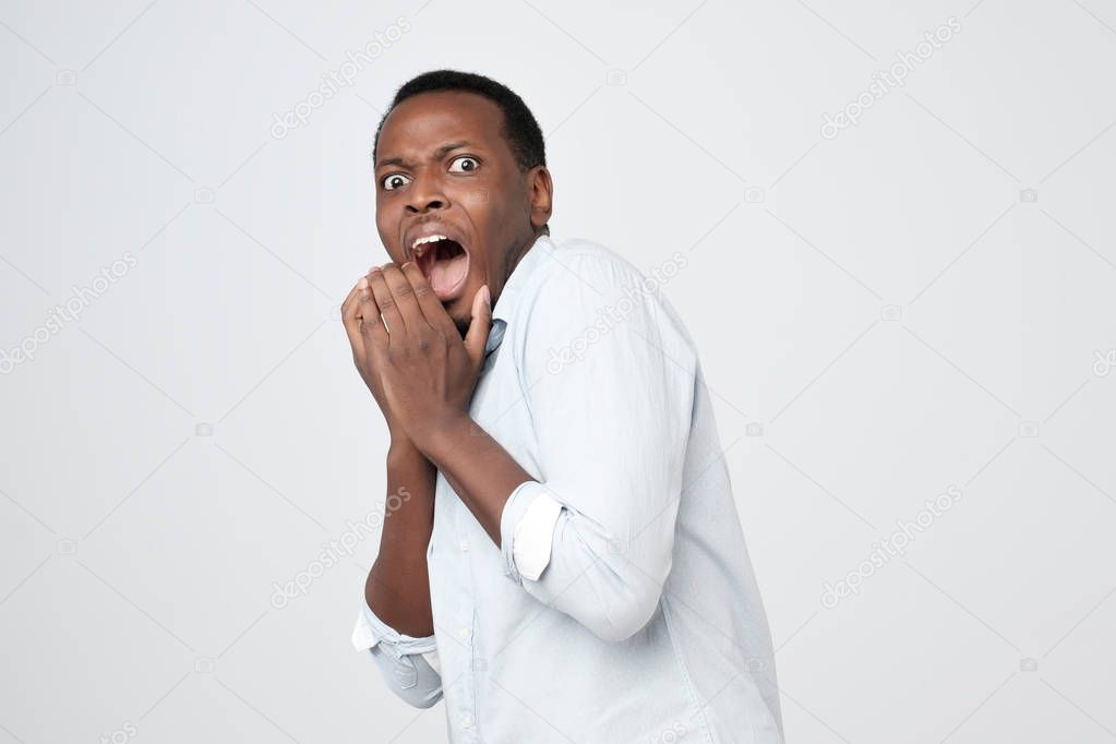 Puzzled african man isolated on grey background dressed in white shirt, covering mouth with hands