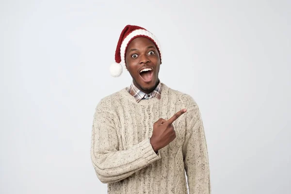 Handsome african man in red christmas cap pointing fingers and looking at camera. — Stock Photo, Image