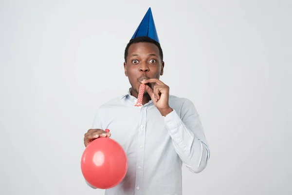Africký mladík baví na strany na sobě modrou košili a prázdninový klobouk, foukání roh stran. — Stock fotografie