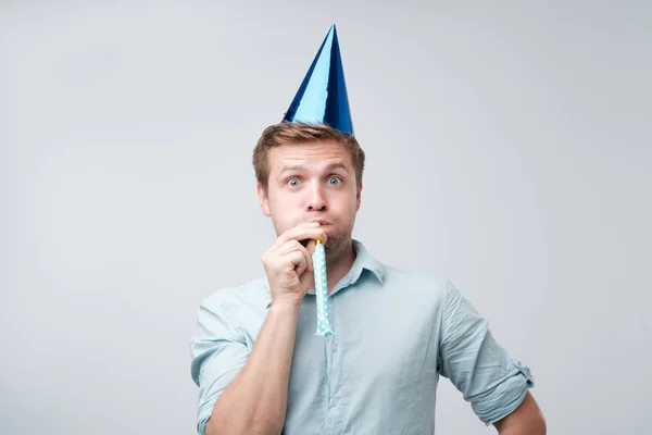Cheerful young man having fun on party wearing blue denim shirt and holiday hat, blowing party horn. — Stock Photo, Image