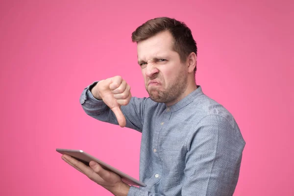 Joven hipster europeo haciendo mala señal mostrando el pulgar hacia abajo sosteniendo la tableta en las manos . —  Fotos de Stock