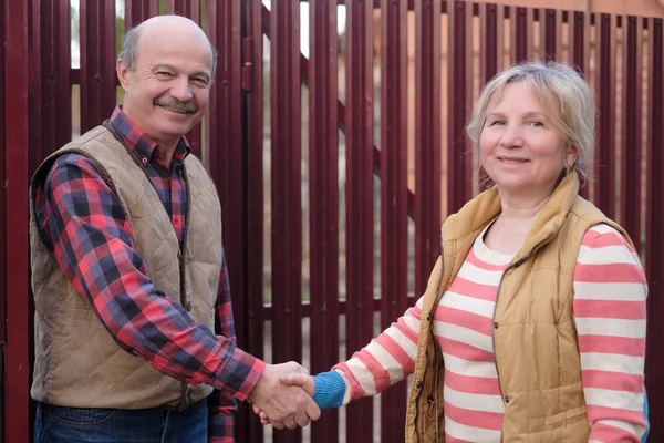 Two senior neighbors takling to each other on sunny day near fence.