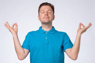 Mavi t-shirt genç yakışıklı adam rahatlatıcı ve gözleri meditasyon jest yapıyor kapalı gülümseyerek