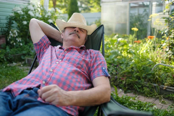 Homme hispanique senior dans le chapeau assis appuyé sur la chaise dormant dans le jardin de fleurs d'été extérieur — Photo