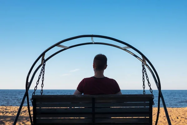 Hombre sentado en columpio en la playa de arena y mirando al mar — Foto de Stock