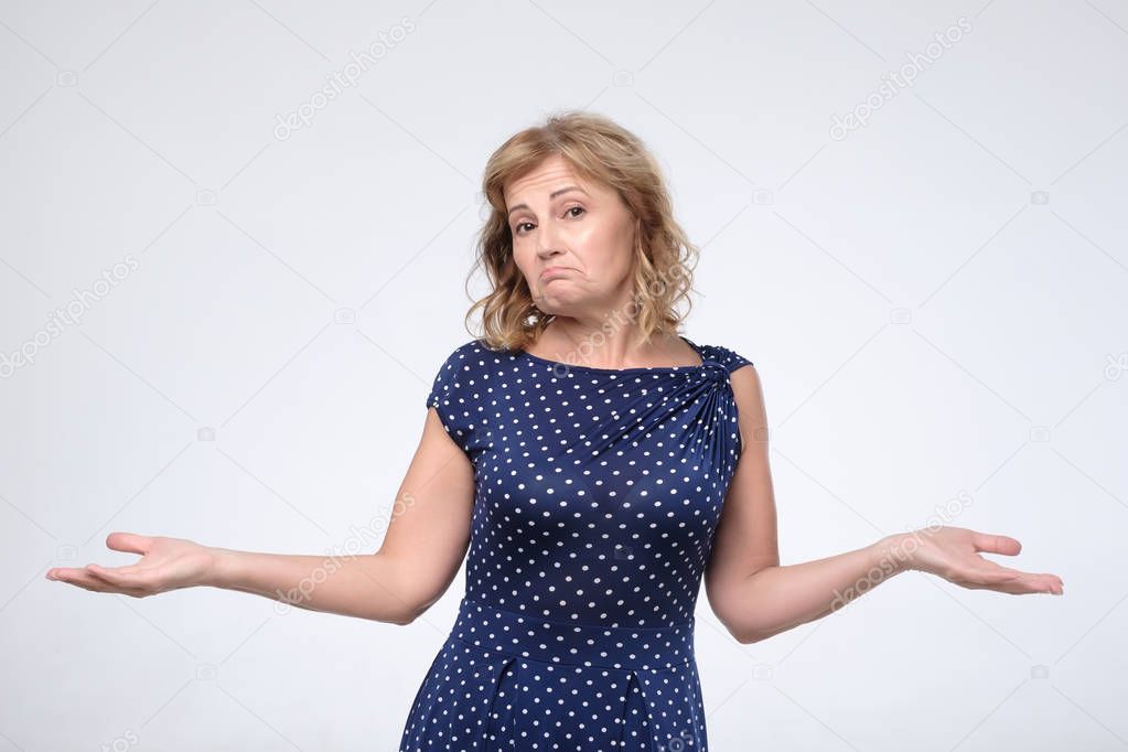 Puzzled mature female in blue dress shrugs shoulders as does not know answer,