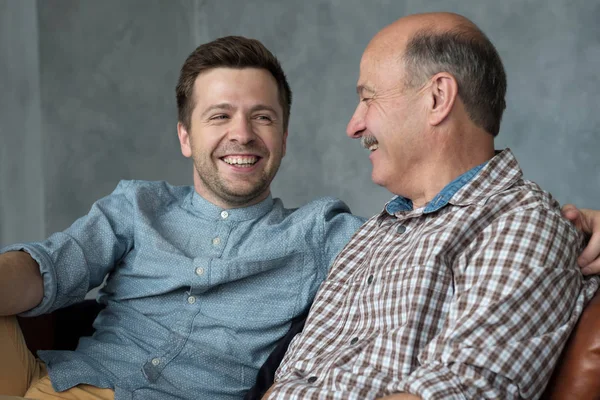 Sorrindo pai sênior sentar no sofá conversa bate-papo com filho milenar passar tempo em casa juntos — Fotografia de Stock