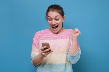 Mavi duvar arka planında cep telefonu kullanarak izole edilmiş mutlu ve iyimser genç bir kadının görüntüsü.