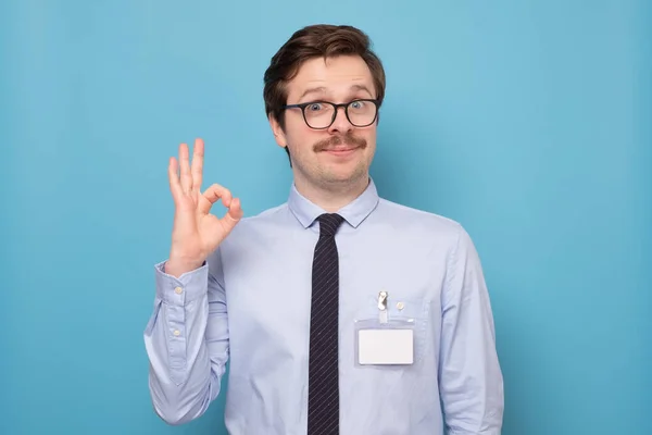 Happy young man in shirt and tie gesturing OK sign on blue wall.