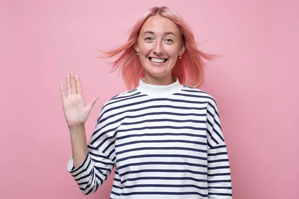 Friendly looking young woman with pink dyed hair smiling happily, saying Hello
