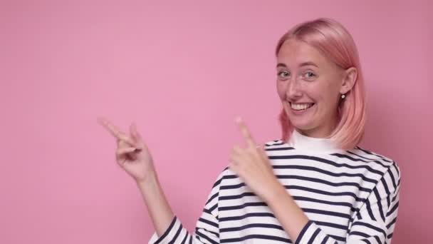 Pozitív fiatal nő festett rózsaszín hajjal vidáman mosolyog és mutatva mutatóujjával el — Stock videók