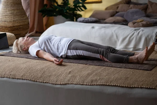 Senior vrouw uit te werken thuis, doen yoga oefening liggend in Shavasana of lichaam of dood lichaam houding — Stockfoto