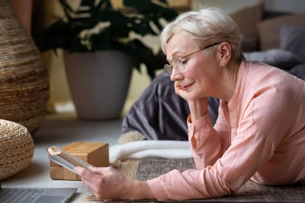 Senior kvinna i glasögon läser en bok hemma. — Stockfoto