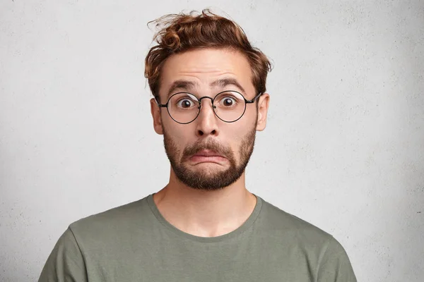 Scared emotional bearded man worries before important event, stares through glass spectacles. Unshaven handsome male looks in astonishment, isolated over white background.